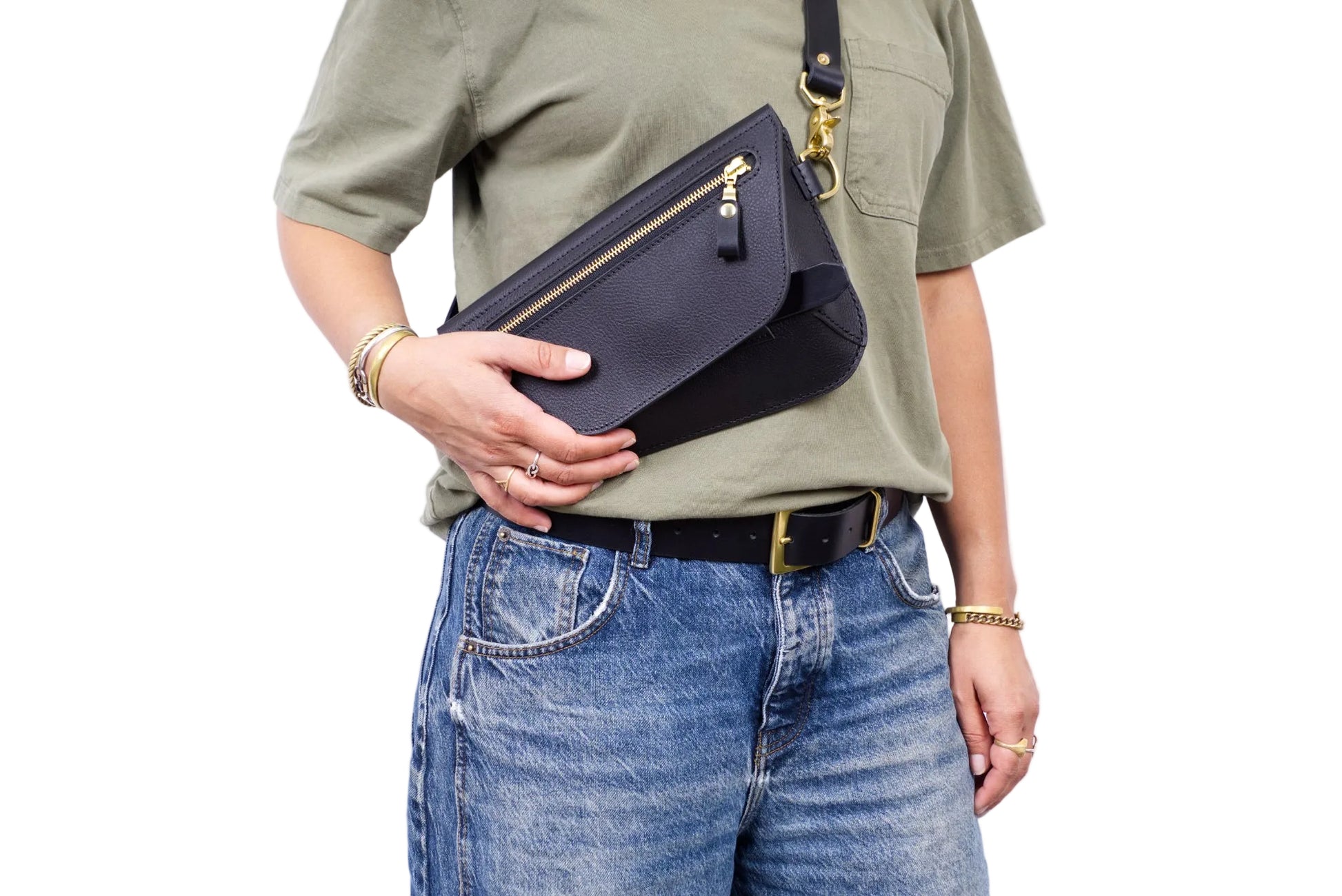 A person wearing an olive green t-shirt and blue jeans holds a black leather crossbody bag with gold-tone hardware against a plain white background.